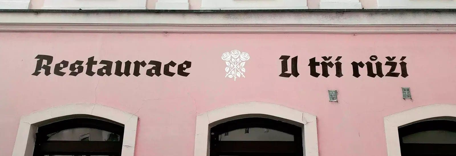 Nápis Restaurace U tří růží lomeným písmem na růžové fasádě